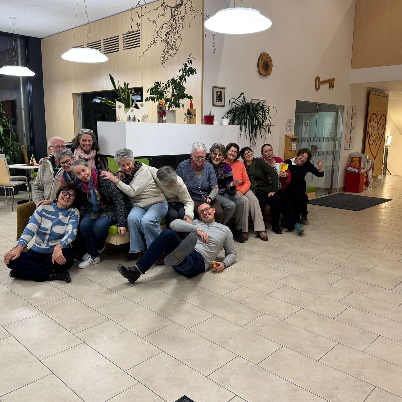 Eine Gruppe von Menschen sitzt und liegt auf einer Bank in einem Aufenthaltsbereich und posiert für ein Foto. Einige tragen Brillen und Turnschuhe, während einer ein Spielzeug hält. Blumentöpfe und Lampen schmücken den Raum.