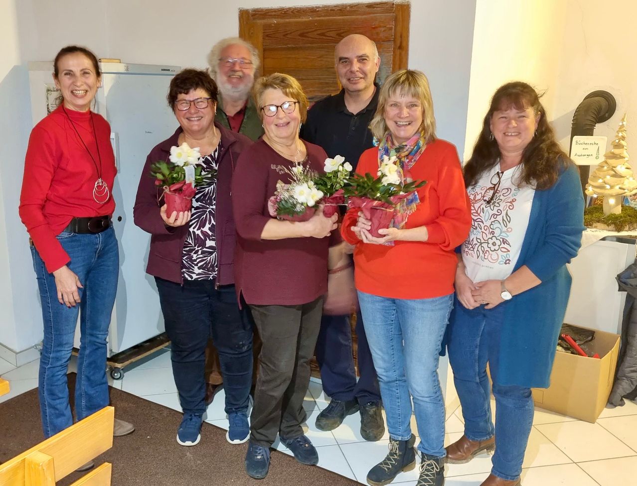 Eine Gruppe von Menschen steht in einem Raum zusammen. Sie halten Blumenvasen und lächeln. Der Boden ist gefliest und die Wand ist weiß gestrichen. Eine Tür befindet sich hinter ihnen.