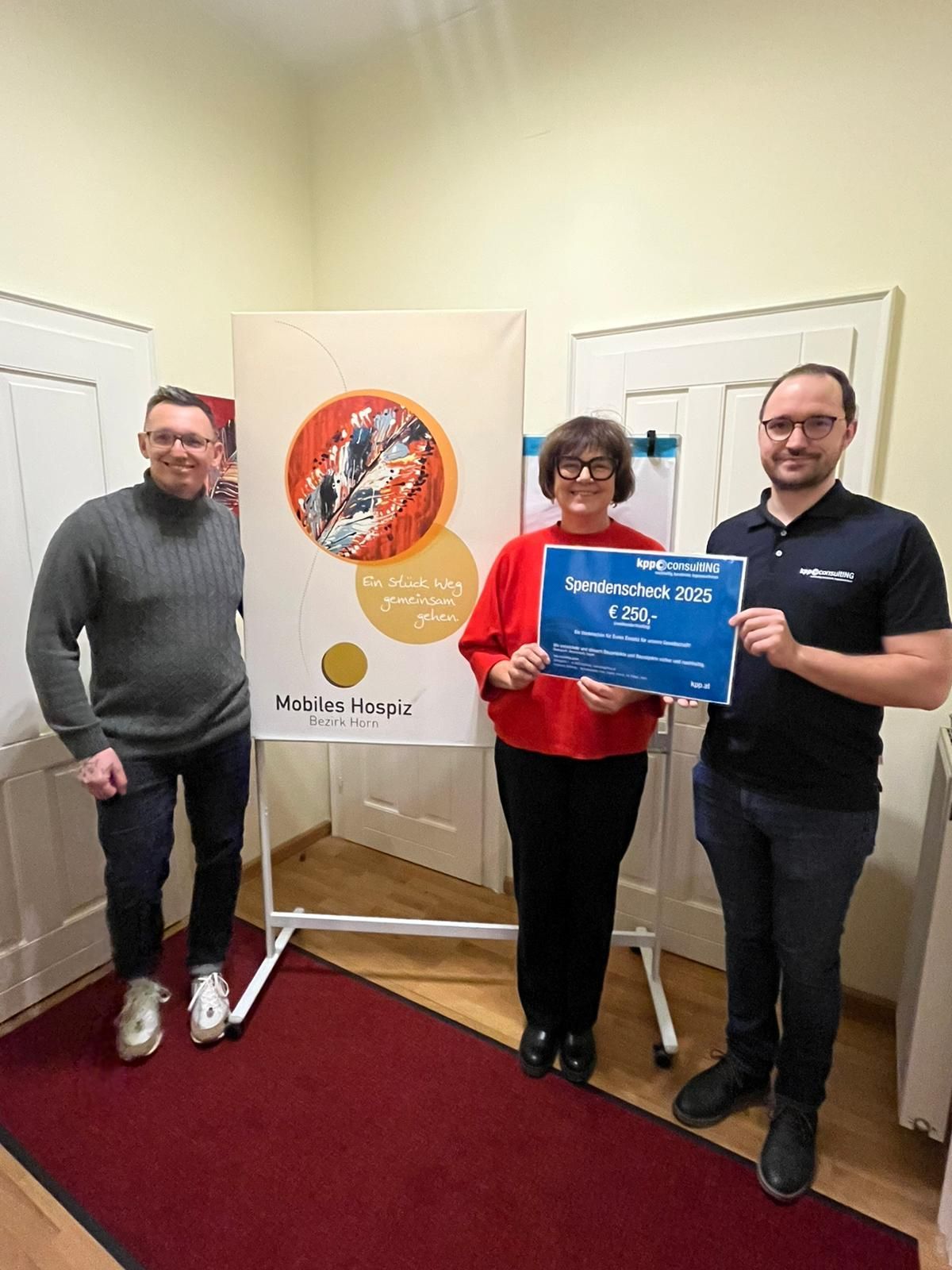 Drei Personen stehen in einem Raum, lächeln und halten ein Banner und einen Spendenscheck. Die Frau hält den Scheck, während der Mann neben ihr lächelt. Hinter ihnen ist ein Banner mit einem farbenfrohen Design.