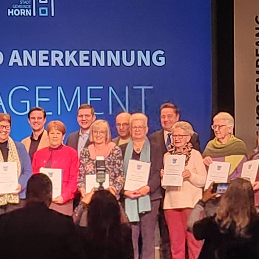 Eine Gruppe von Menschen steht mit Urkunden auf der Bühne, lächelt für die Kamera, vor einem blauen Hintergrund mit der Aufschrift 'ANerkennung Management'.