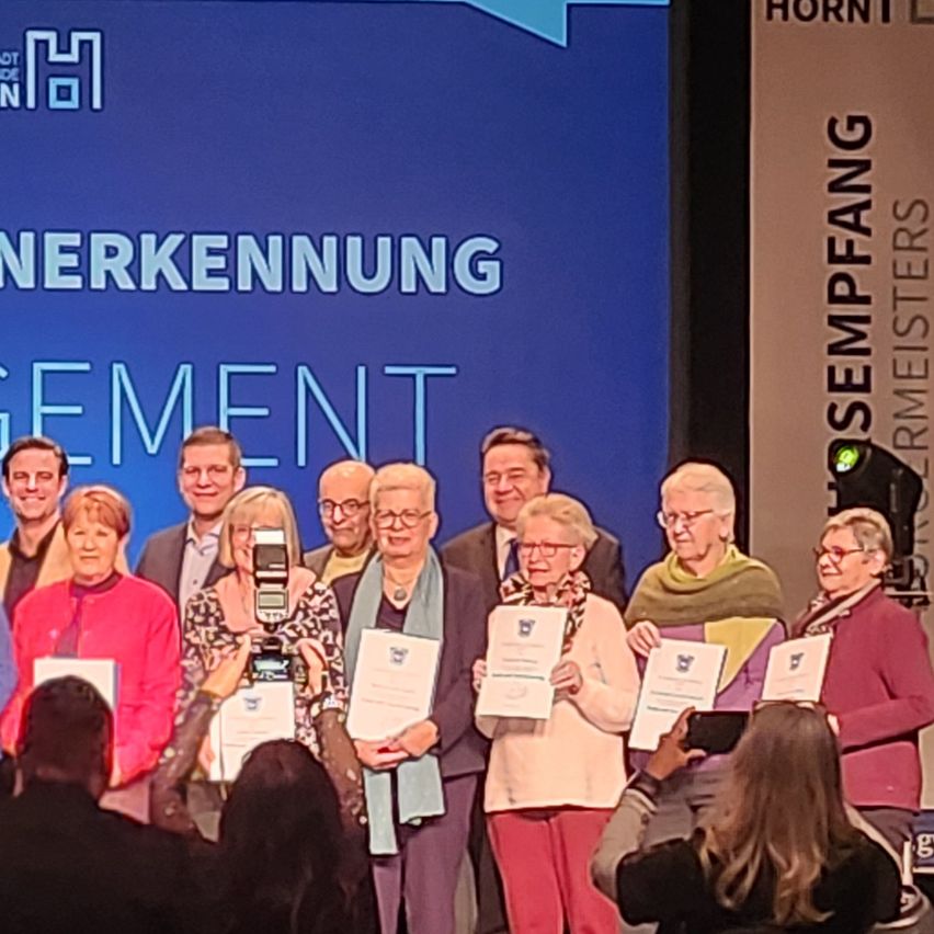 Eine Gruppe von Menschen, darunter Männer und Frauen, steht vor einem blauen Hintergrund, hält Urkunden in den Händen und posiert für ein Foto. Sie tragen alle Brillen und einige haben Schals um. Rechts befindet sich eine Lampe.