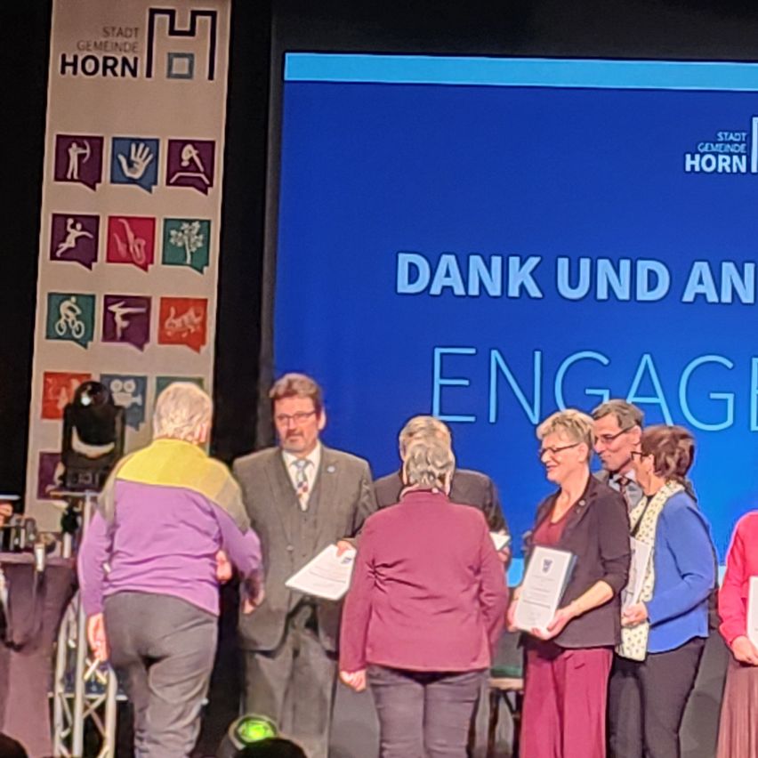 Eine Gruppe von Menschen steht auf der Bühne und erhält Urkunden. Ein blauer Bildschirm hinter ihnen zeigt 'DANK UND AN ENGAG'. Ein Banner mit verschiedenen Symbolen und dem Wort 'HORN' ist links.
