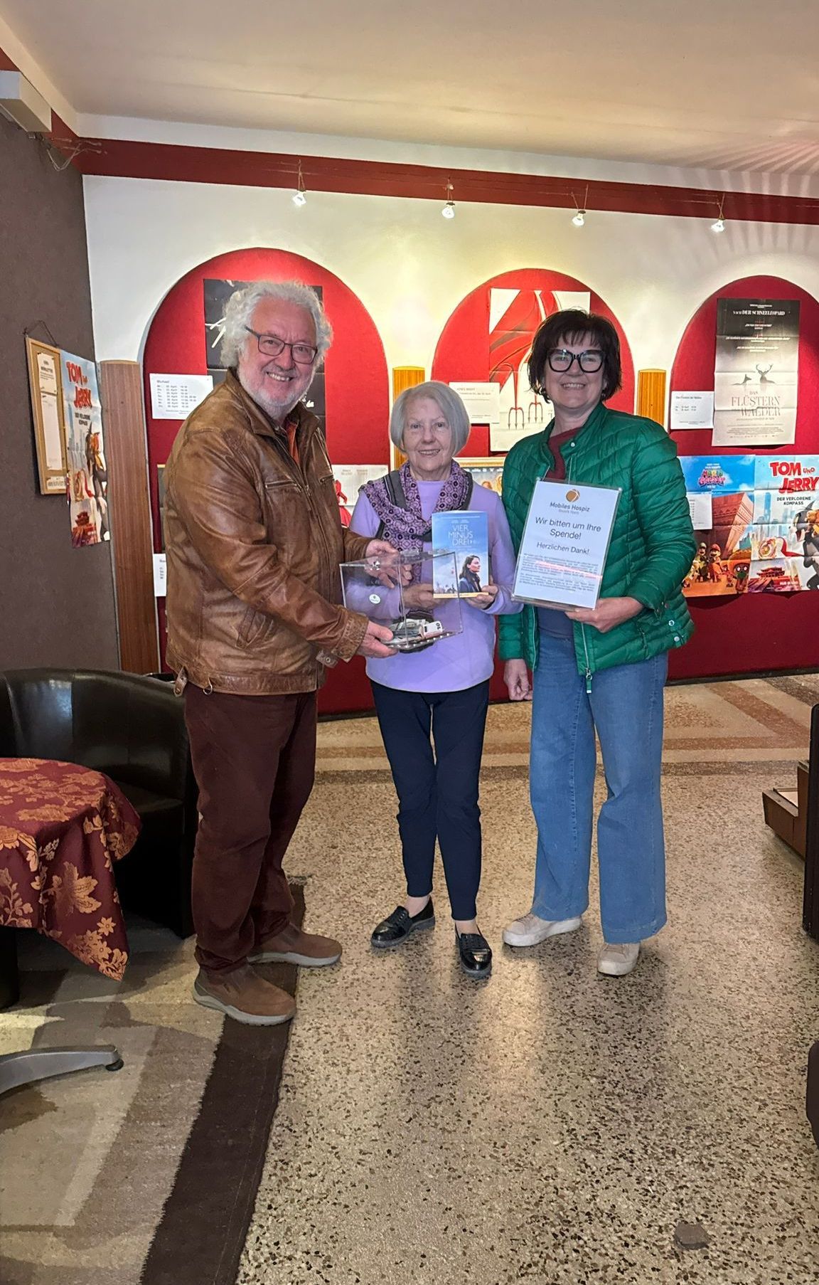 Drei Personen posieren für ein Bild und halten Bücher und Urkunden in der Hand, in einem Raum mit roten Bögen und Filmplakaten im Hintergrund.