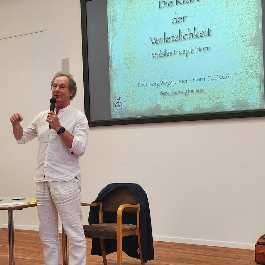 Ein Mann spricht in ein Mikrofon in einem Raum. Ein großer Bildschirm hinter ihm zeigt eine Folie mit deutschem Text über eine Veranstaltung im Mobiles Hospiz Horn.