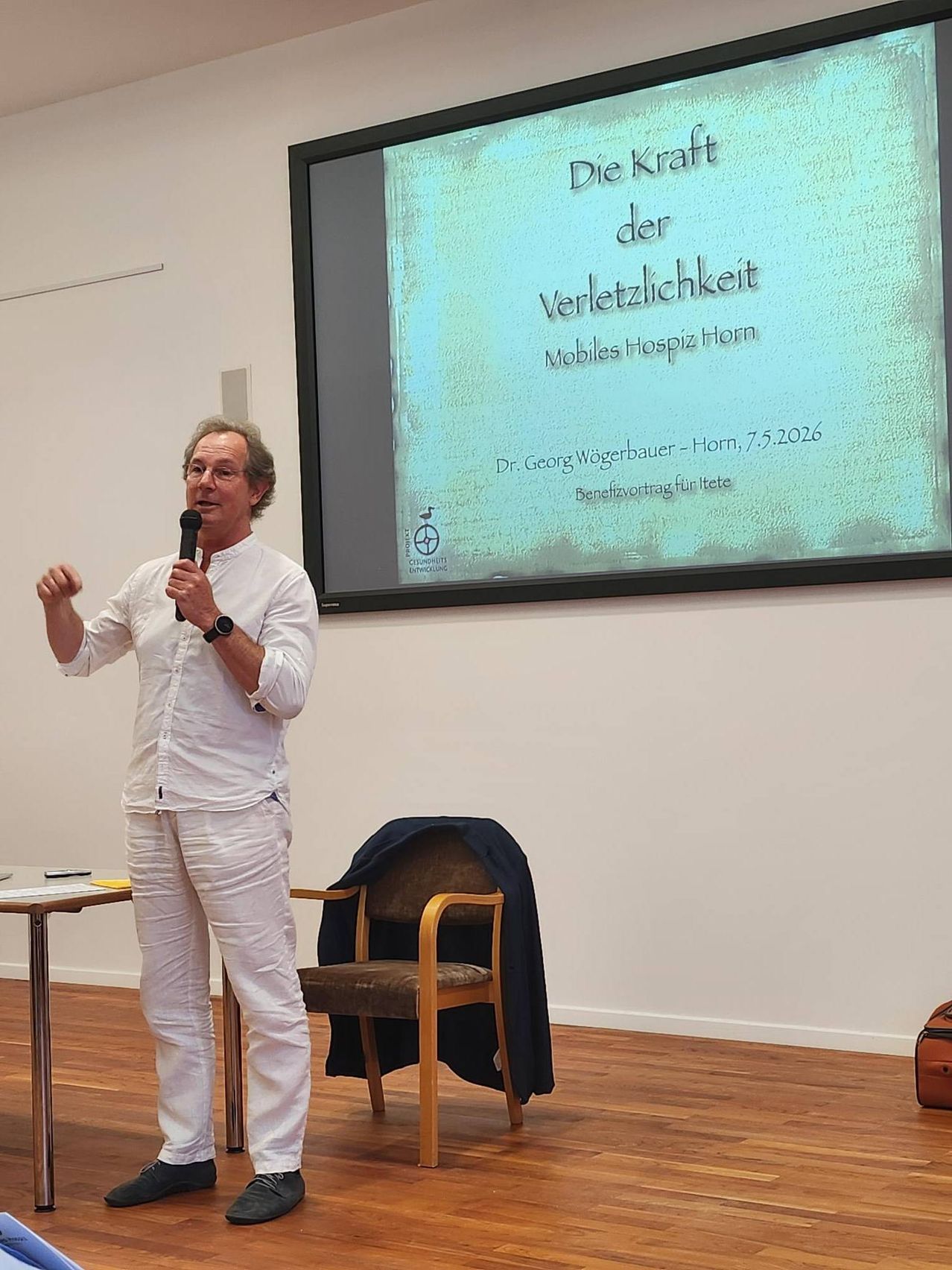 Ein Mann spricht in ein Mikrofon in einem Raum. Ein großer Bildschirm hinter ihm zeigt eine Folie mit deutschem Text über eine Veranstaltung im Mobiles Hospiz Horn.