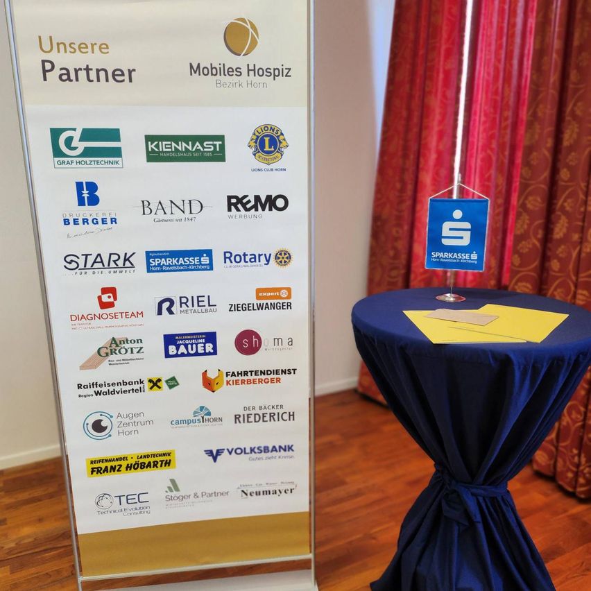 Ein Schild zeigt verschiedene Firmenlogos und ein Partnerschaftsbanner. Es steht vor einer Kulisse mit einem blauen Tischtuch und einem runden Tisch.