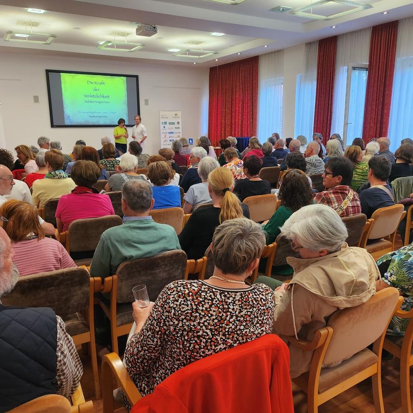 Ein Raum voller Menschen, die eine Präsentation verfolgen, zwei Personen stehen vor einer Projektionsleinwand, eine vor der anderen.