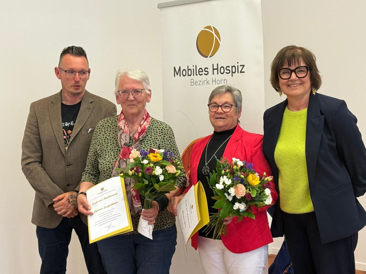 Vier Personen stehen vor einem weißen Banner mit dem Text 'Mobiles Hospiz'. Drei Frauen halten Blumen und Urkunden, während ein Mann hinter ihnen steht. Die Frauen tragen Brillen und Jacken, eine hat eine Halskette. Das Banner zeigt ein Basketball-Logo.