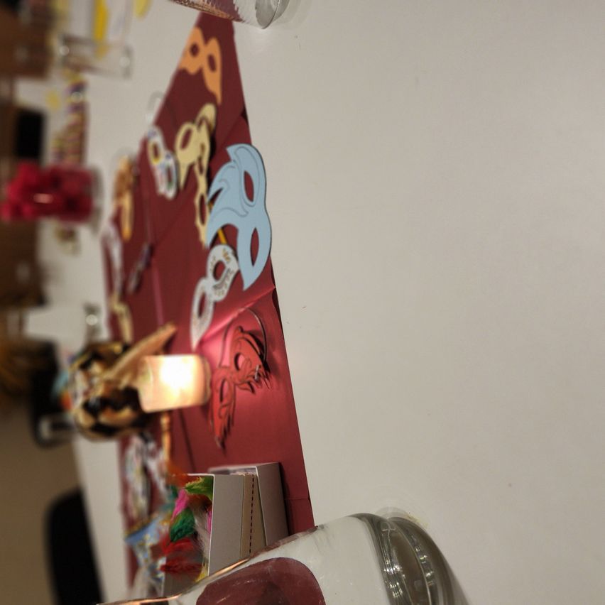 Ein Tisch mit rotem Hintergrund zeigt verschiedene farbenfrohe Dekorationen. Eine Lampe ist beleuchtet, und es hängen mehrere Masken und Bänder an der Wand.