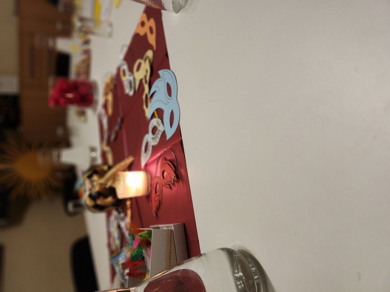 Ein Tisch mit rotem Hintergrund zeigt verschiedene farbenfrohe Dekorationen. Eine Lampe ist beleuchtet, und es hängen mehrere Masken und Bänder an der Wand.