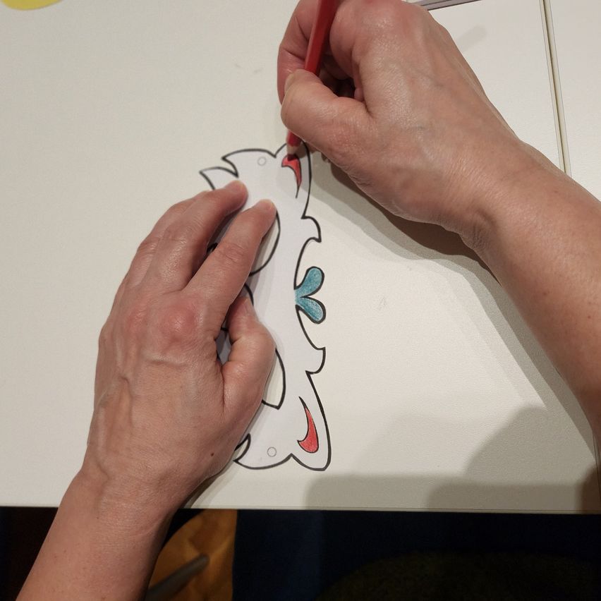 Eine Person zeichnet mit einem roten Stift auf ein Blatt Papier, das sie mit beiden Händen auf einem weißen Tisch hält. Das Papier zeigt eine Zeichnung eines Vogels.