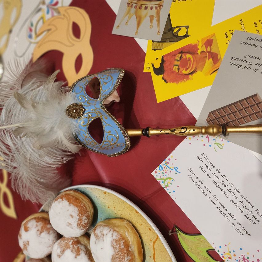 Auf einem Tisch sind eine blau-goldene Maske, ein weißer Federfächer und gelbe Karten zu sehen. Ein roter Tischläufer mit einem Teller mit Puderzucker-Donuts ist ebenfalls vorhanden.