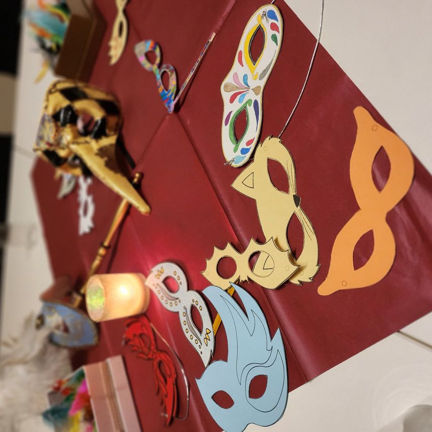 Eine Vielzahl bunter Masken ist auf einer roten Oberfläche ausgestellt. Darunter sind eine goldene Maske, eine blaue Maske und eine orange Maske. Es gibt auch eine Kerze.