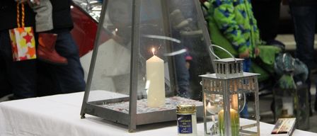 Ein Kerzenarrangement mit einer Laterne steht auf einem weißen Tisch mit Menschen im Hintergrund, darunter ein Junge in einer grünen Jacke.