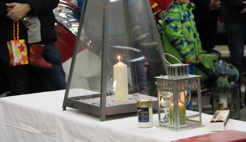 Ein Kerzenarrangement mit einer Laterne steht auf einem weißen Tisch mit Menschen im Hintergrund, darunter ein Junge in einer grünen Jacke.