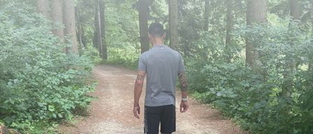 A man walks along a forest path, wearing gray t-shirt and black shorts. Text reads 'Tommy Fit' with a message about active living with Parkinson. Website www.tommyfit.at is visible.