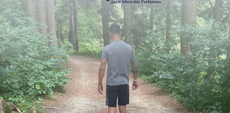 Ein Mann geht einen Waldweg entlang, trägt ein graues T-Shirt und schwarze Shorts. Der Text 'Tommy Fit' enthält eine Botschaft über aktives Leben mit Parkinson. Die Website www.tommyfit.at ist sichtbar.
