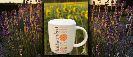 Eine weiße Kaffeetasse mit den Worten 'Ruhe Kraft Freude' in einem Lavendelfeld mit Bäumen im Hintergrund.