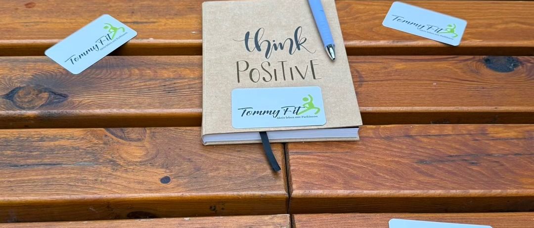 A notebook with the words 'Think Positive' is placed on a wooden bench. A blue pen is resting on the notebook. Four Tommy Fit cards are scattered around the bench.