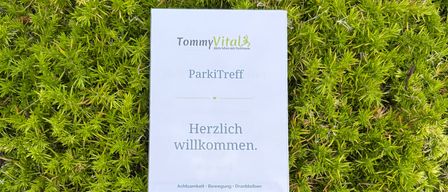 Ein weißes Schild mit grünem Text, das 'TommyVital Aktiv leben mit Parkinson' und 'ParkiTreff Herzlich willkommen' besagt, liegt auf einer grünen moosigen Oberfläche.