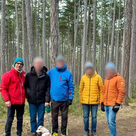 Fünf Personen in Winterkleidung stehen in einem Wald. Einer hat einen Hund an der Leine. Sie alle schauen geradeaus, ihre Gesichter sind verschwommen.