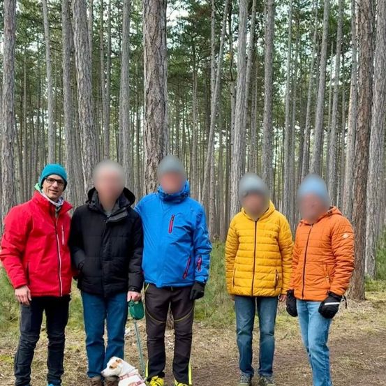 Fünf Personen in Winterkleidung stehen in einem Wald. Einer hat einen Hund an der Leine. Sie alle schauen geradeaus, ihre Gesichter sind verschwommen.