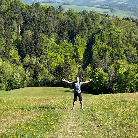 Ein Mann steht mit ausgestreckten Armen in einem Grasfeld, umgeben von einem Wald und Hügeln im Hintergrund.