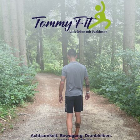 Ein Mann mit Tätowierungen an Armen und Beinen geht auf einem Pfad im Wald, trägt ein graues Shirt und schwarze Shorts. Der Pfad ist von hohen Bäumen und grünen Pflanzen umgeben. Die Worte 'Tommy Fit' sind oben angezeigt.