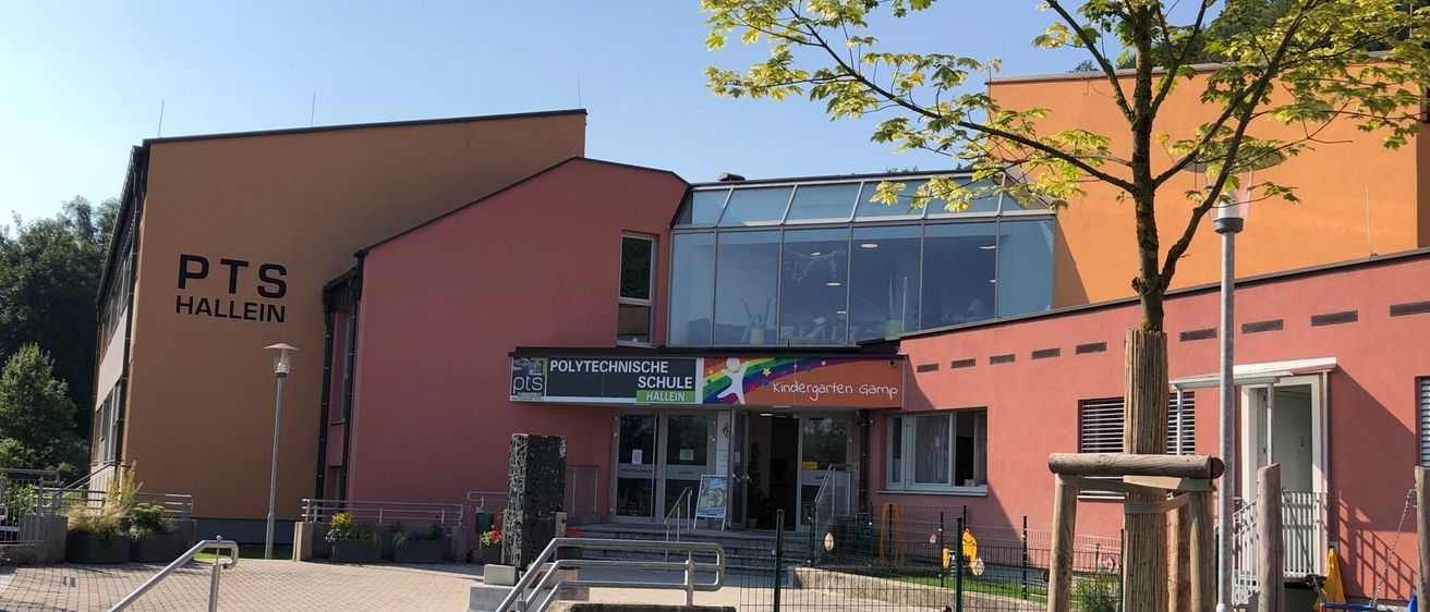 Ein Schulgebäude mit roter Fassade und Glasfenstern. Es hat ein Schild mit der Aufschrift 'PTS Hallein'. Vor dem Gebäude befindet sich ein kleiner Garten und ein Spielplatz.