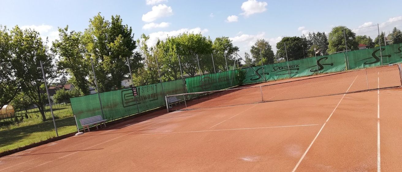 Ein leerer Tennisplatz mit einem grünen Zaun, Bänken und Bäumen unter einem blauen Himmel mit einigen Wolken.