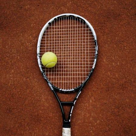 Ein Tennisschläger mit einem Tennisball auf den Saiten liegt auf einem braunen Sandplatz.