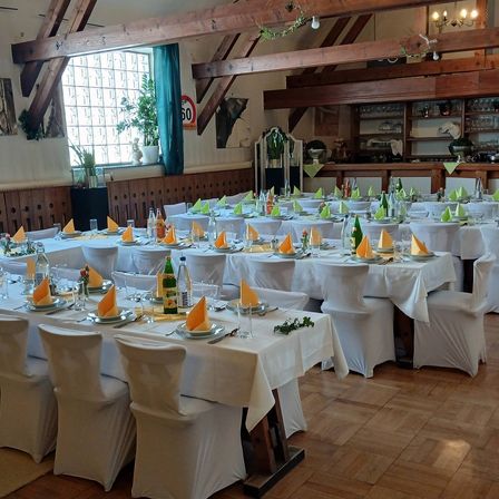 Ein Indoor-Bankettsaal mit mehreren Tischen, die mit weißen Tischdecken bedeckt sind. Jeder Tisch hat Teller, Gläser und Servietten. Weinflaschen und Blumen sind ebenfalls vorhanden. Holzbalken und ein Geschwindigkeitsschild sind sichtbar.