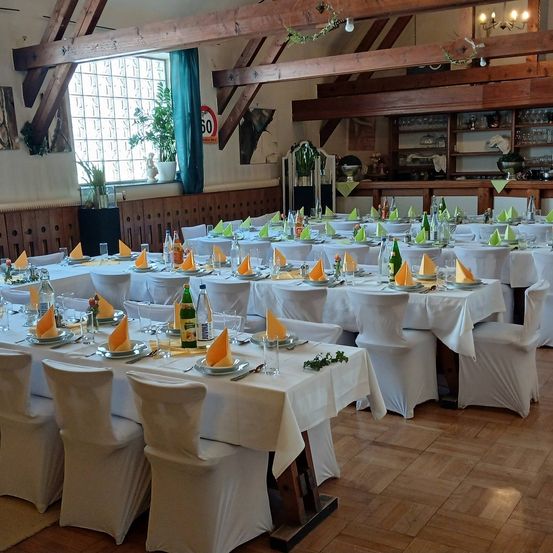 Ein Indoor-Bankettsaal mit mehreren Tischen, die mit weißen Tischdecken bedeckt sind. Jeder Tisch hat Teller, Gläser und Servietten. Weinflaschen und Blumen sind ebenfalls vorhanden. Holzbalken und ein Geschwindigkeitsschild sind sichtbar.