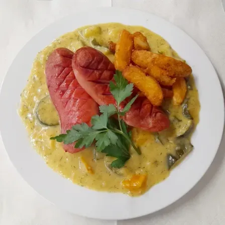 Ein weißer Teller mit einem cremigen Gericht und zwei Scheiben Hot Dogs, garniert mit Petersilie, serviert mit einer Beilage aus gebratenen Kartoffeln und Zucchini auf einem weißen Tuch.