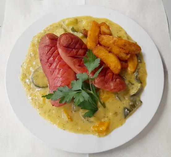 Ein weißer Teller mit einem cremigen Gericht und zwei Scheiben Hot Dogs, garniert mit Petersilie, serviert mit einer Beilage aus gebratenen Kartoffeln und Zucchini auf einem weißen Tuch.