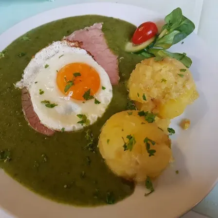 Ein Teller mit grüner Soße, zwei gebratenen Eiern, einem Stück Schinken, Kartoffelpüree und einer Tomate obenauf.