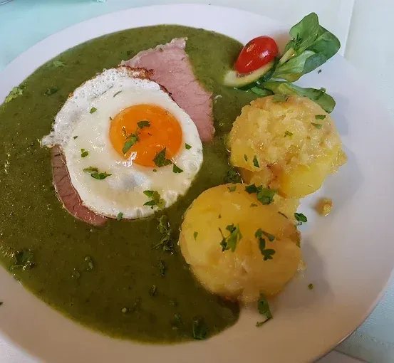 Ein Teller mit grüner Soße, zwei gebratenen Eiern, einem Stück Schinken, Kartoffelpüree und einer Tomate obenauf.