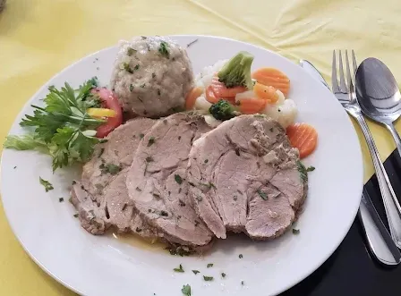 Ein weißer Teller mit geschnittenem Schweinefleisch, Knödeln und Gemüse. Eine Gabel, ein Messer und ein Löffel sind rechts. Ein Glas und ein roter Platzset sind auf dem Tisch.