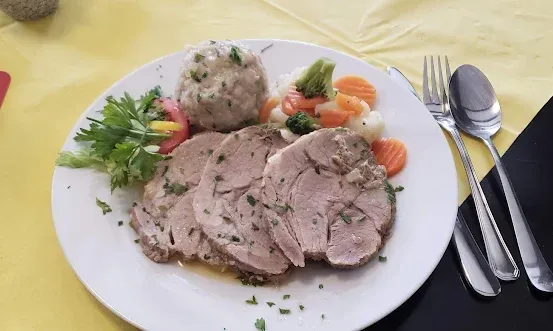 Ein weißer Teller mit geschnittenem Schweinefleisch, Knödeln und Gemüse. Eine Gabel, ein Messer und ein Löffel sind rechts. Ein Glas und ein roter Platzset sind auf dem Tisch.