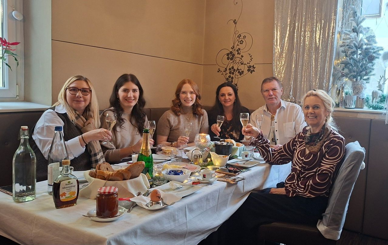 Eine Gruppe von Menschen sitzt um einen Tisch herum, lächelt und hält Gläser mit Champagner. Der Tisch ist mit Tellern, Schüsseln, einer Kerze und einer Flasche gedeckt. Sie tragen Winterkleidung.