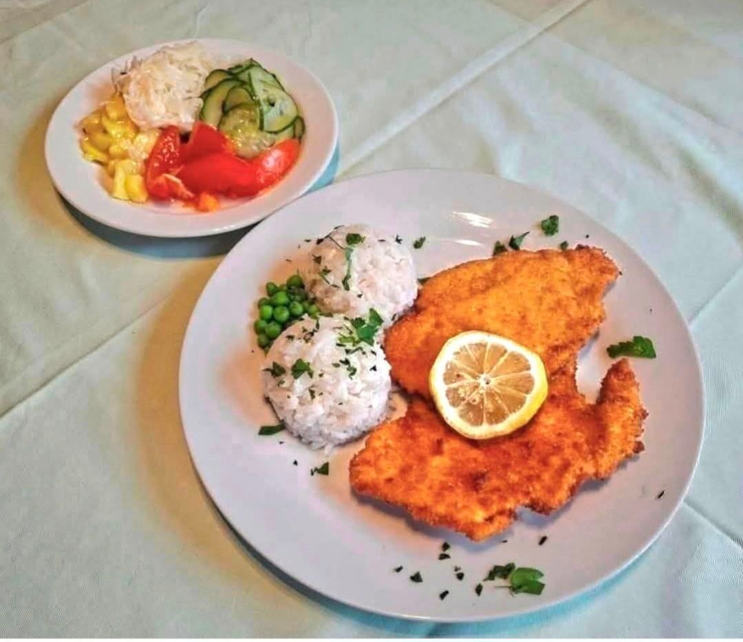 Ein Teller mit gebratenem Hähnchen mit Reis und Erbsen und einer Zitronenscheibe. Ein weiterer Teller enthält Gurken, Tomaten und Karotten. Beide Teller stehen auf einem Tisch.