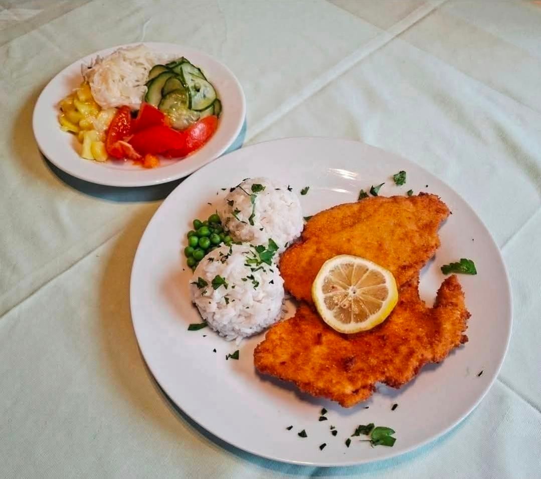 Zwei Teller mit Essen auf einem Tisch. Ein Teller hat panierte Hähnchenschnitzel mit Reis und Erbsen. Der andere Teller hat Gemüse.