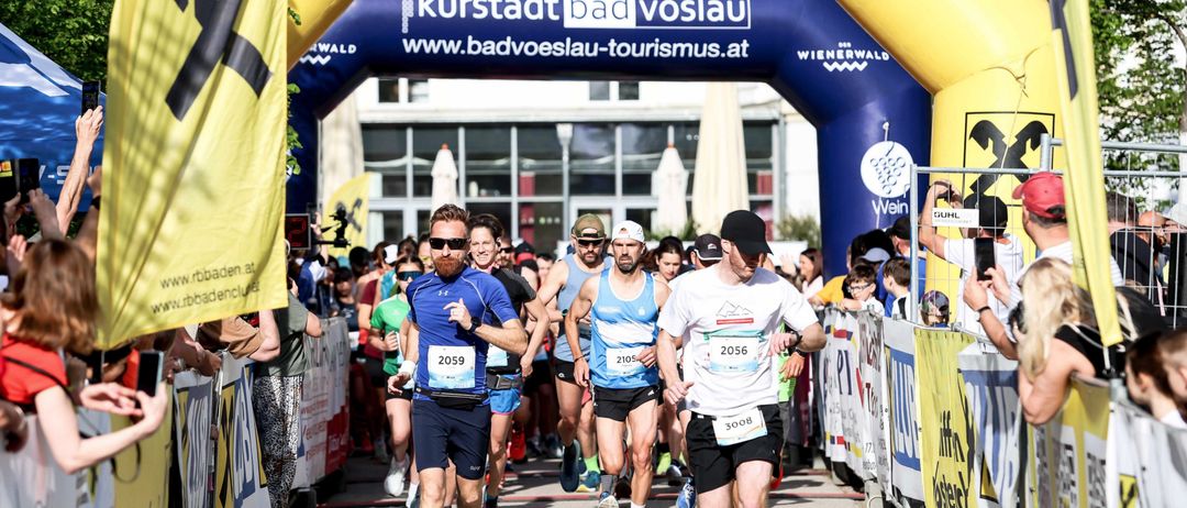 Läufer durchlaufen einen gelben Bogen, der mit Raiffeisenbank Baden am Bad Vöslau-Marathon gekennzeichnet ist. Zuschauer ermutigen die Läufer an der Seite.