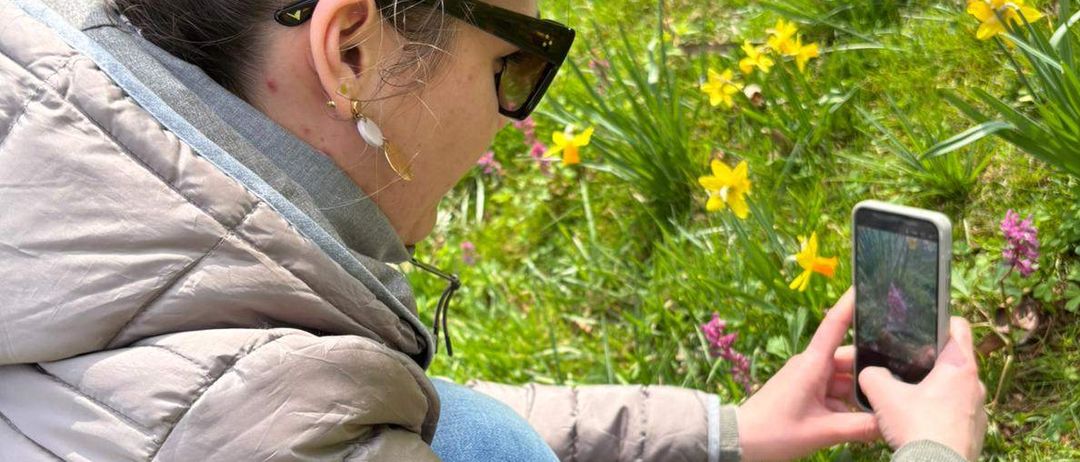 Eine Frau in einer flauschigen Jacke mit Sonnenbrille macht mit ihrem Handy ein Foto, während sie in einem Feld mit gelben Blumen sitzt.