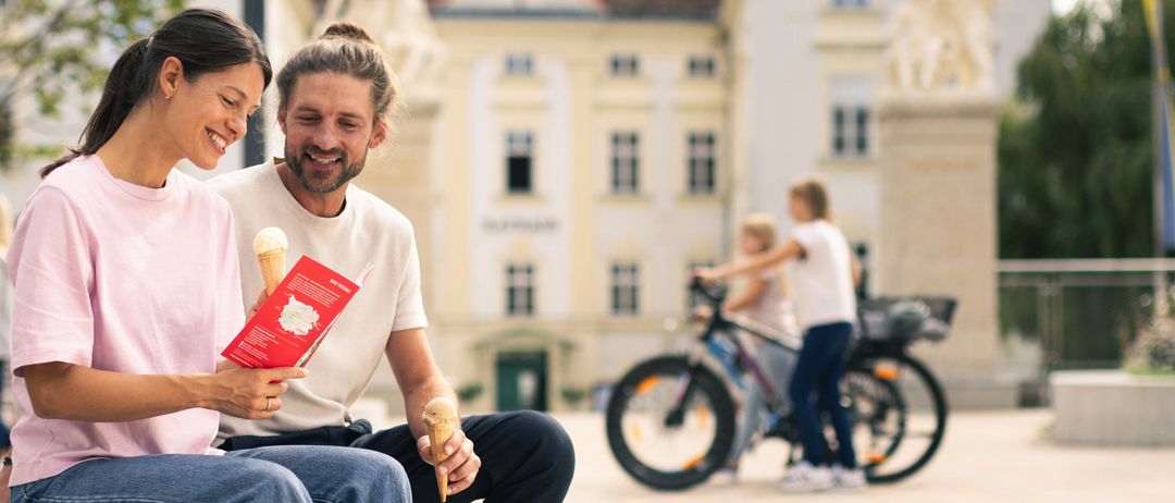 Ein Paar sitzt auf einer Bank vor einem historischen Gebäude und genießt Eis. Dahinter fährt eine Person mit einem Fahrrad vorbei. Das Wetter ist sonnig.
