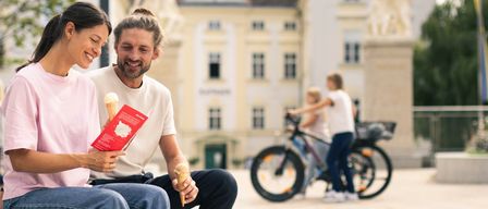 Ein Paar genießt Eis vor einem historischen Gebäude, während ein Kind im Hintergrund mit dem Fahrrad fährt.