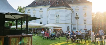 Eine Gruppe von Menschen sitzt an Tischen auf einer Rasenfläche vor einem großen Gebäude mit vielen Fenstern und einem steilen Dach.