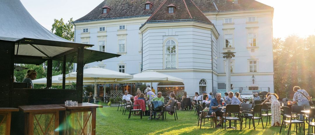 Eine Außengastronomie mit Leuten, die unter Sonnenschirmen an Tischen sitzen, vor einem großen Gebäude mit vielen Fenstern.