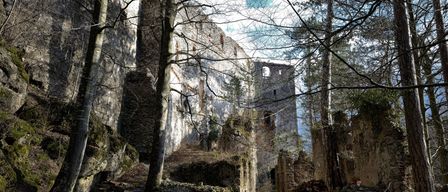 Ruinen einer alten Burg umgeben von Bäumen und Steinmauern. Ein Turm ragt hoch aus den Trümmern. Der Himmel ist klar und blau.