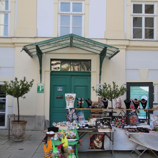 Eine grüne Tür mit einem Vordach befindet sich vor einem Gebäude mit Glasfenstern. An den Seiten stehen Topfpflanzen. Auf dem Bürgersteig gibt es Tische mit verschiedenen Artikeln zum Verkauf.